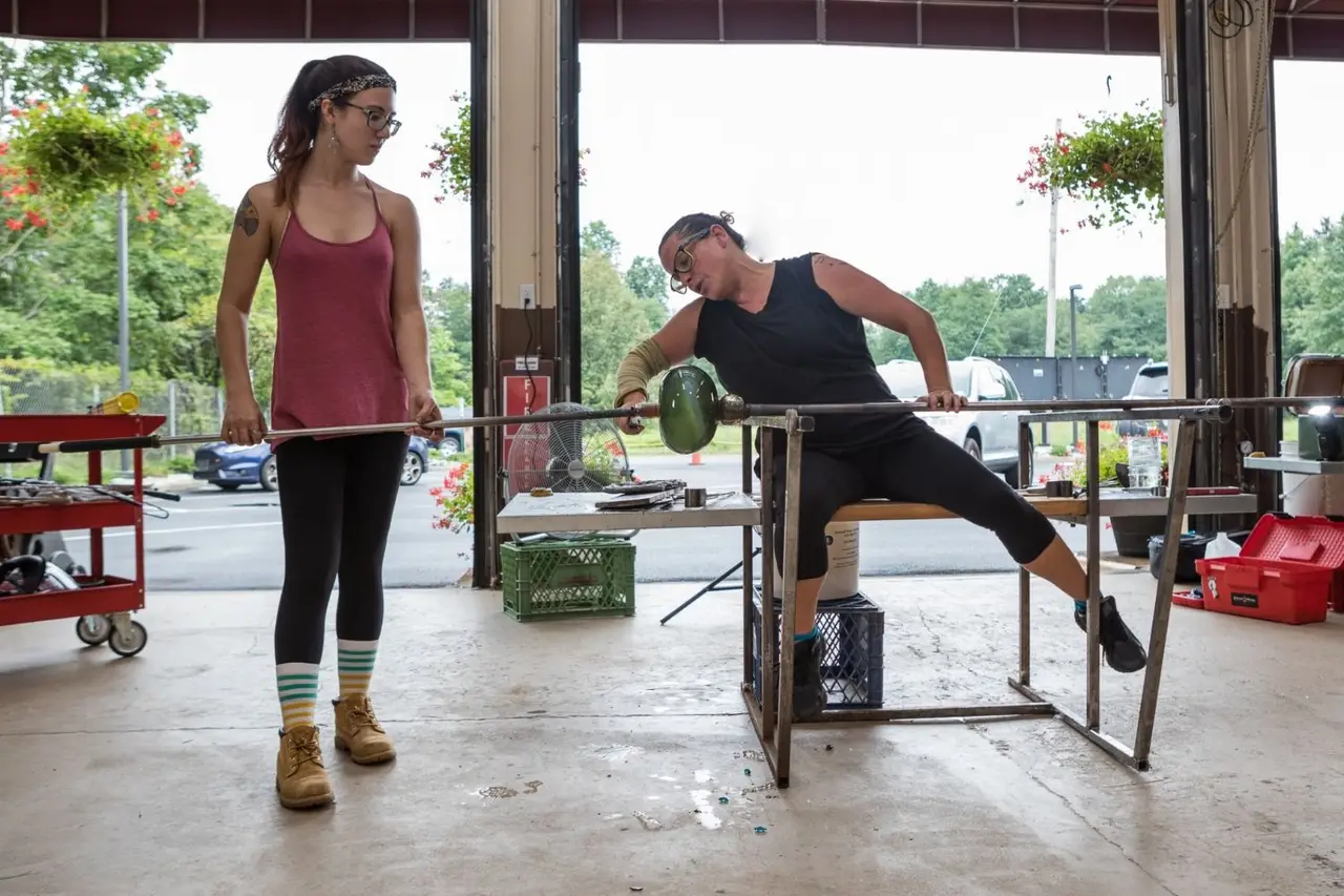 Two glassblowers at work in the hot shop with the garage doors open to the summer