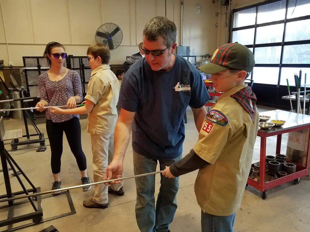A scout in uniform works a pipe of hot glass with an instructor while his troop watches