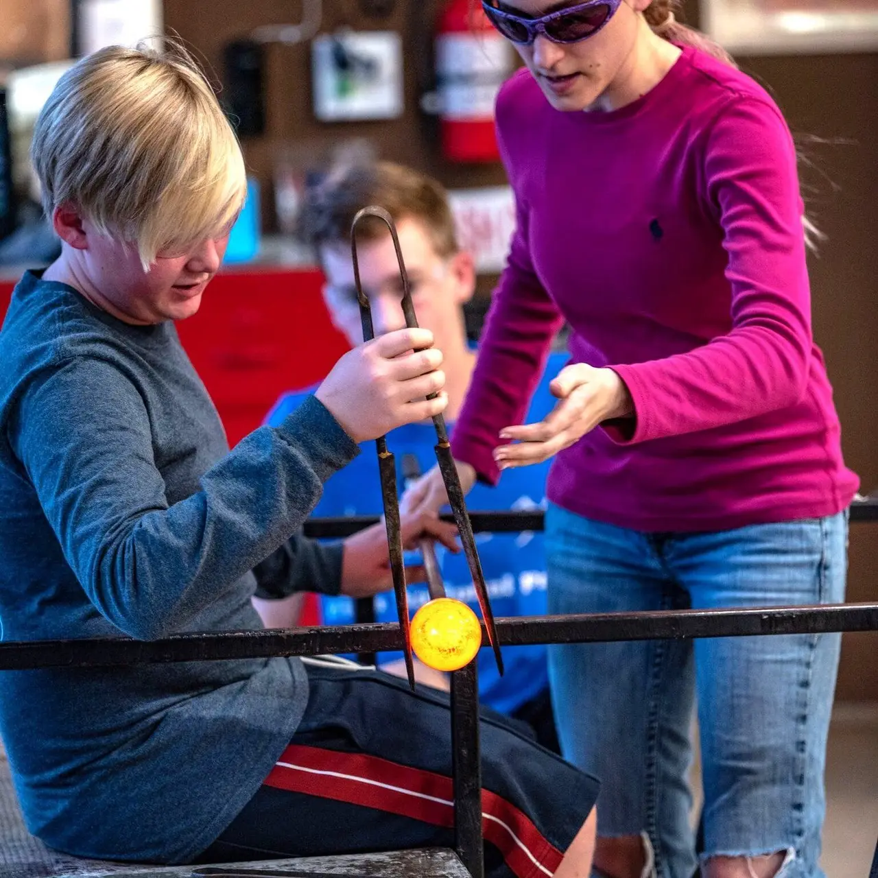 A teenager shapes a glowing piece at the bench with an instructor beside him